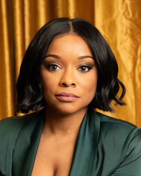 LaToya Lane, seated, in a dark green suit in front of a gold curtain