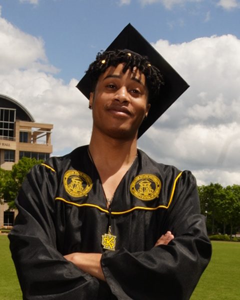 Patrick Arnold in a black graduation cap and gown, arms folded, on a university lawn