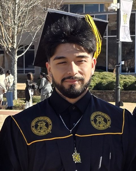 Viz Garcia in a black graduation cap and gown with a gold tassel, outside a university building